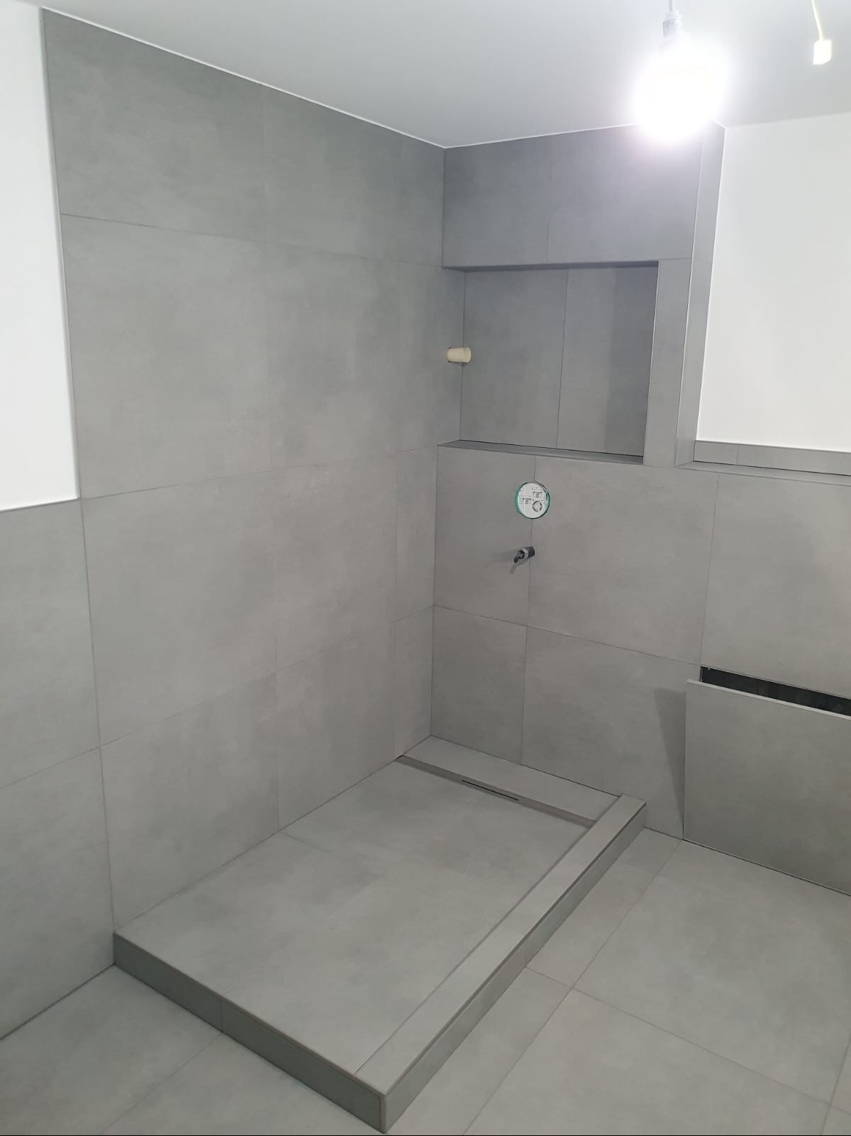 Wykończona, nowoczesna kabina prysznicowa walk-in z szarymi, wielkoformatowymi płytkami, minimalistycznym odpływem liniowym i wnęką w ścianie. Bateria podtynkowa z okrągłą rozetą.