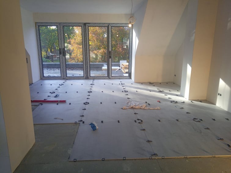 Przygotowanie podłoża pod płytki w jasnym pomieszczeniu z dużymi oknami balkonowymi, widoczne poziomice i system poziomowania płytek.