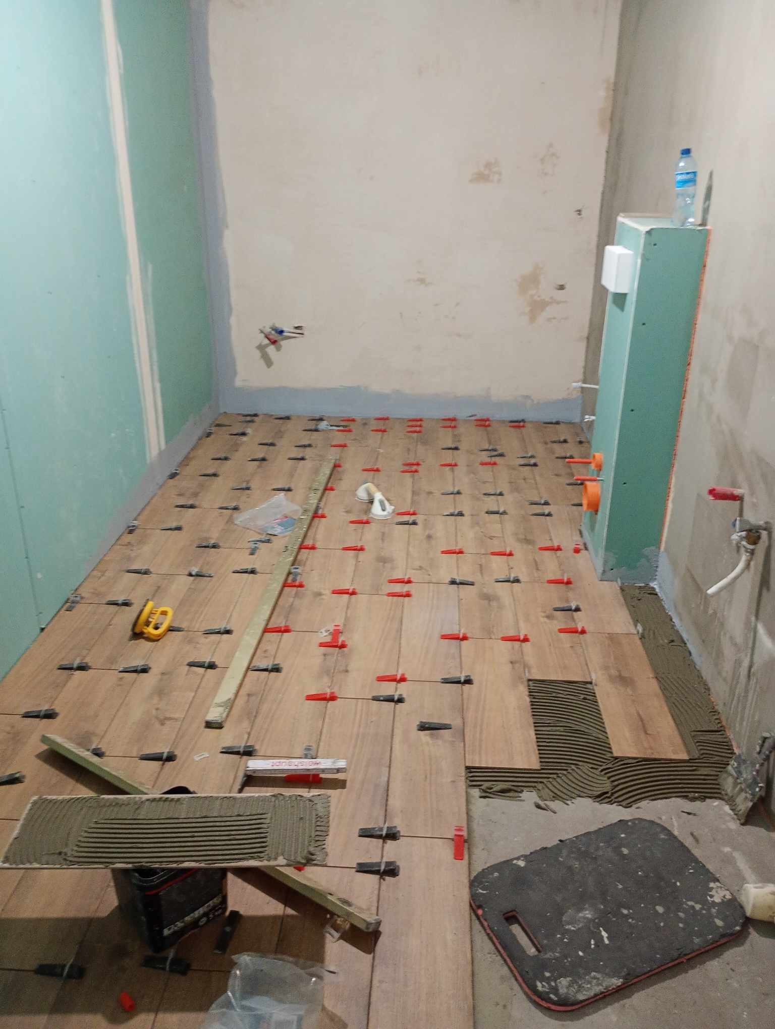 Układanie drewnopodobnych płytek na podłodze w trakcie prac, widoczne poziomice, krzyżyki dystansowe i klej na płytkach, ściany częściowo przygotowane, butelka wody w rogu pomieszczenia.