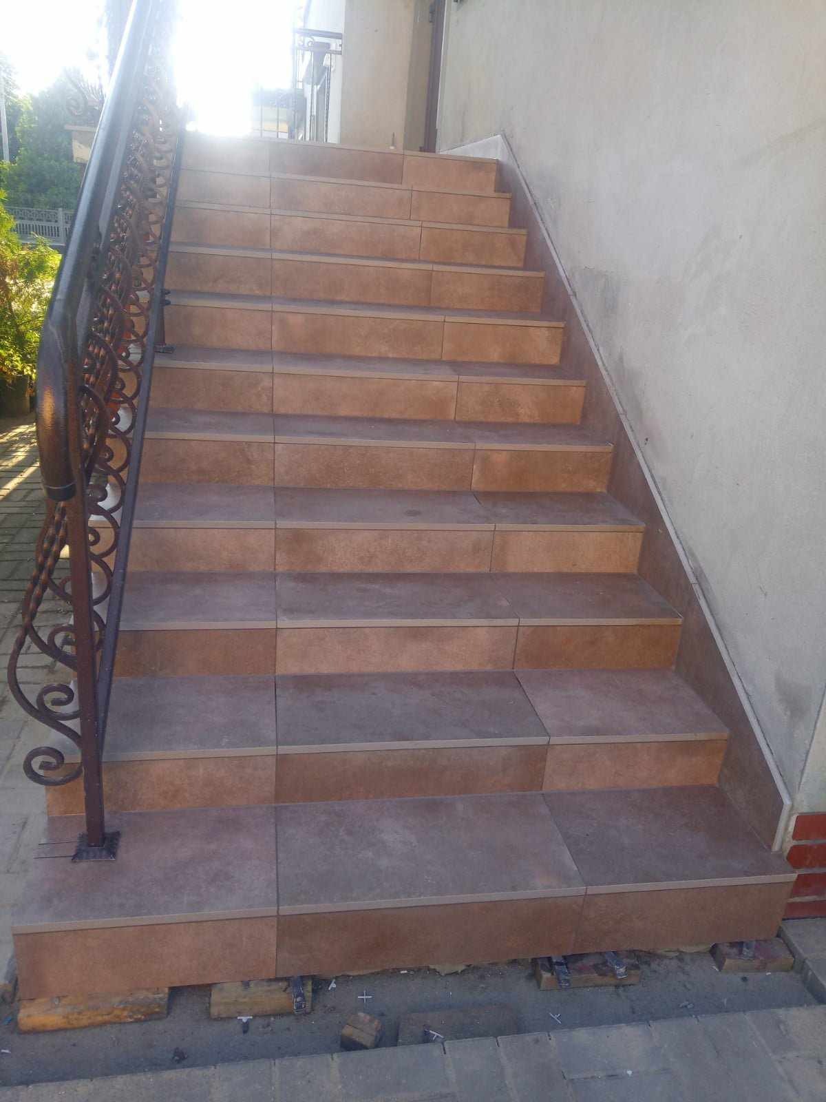 Schody zewnętrzne wyłożone brązowymi płytkami, z ozdobną metalową balustradą, widoczne ślady prac wykończeniowych u podstawy.
