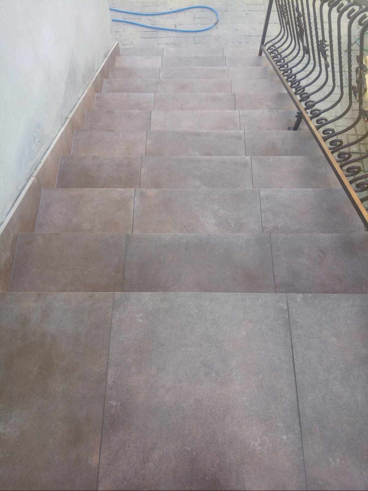 Schody wyłożone szarymi płytkami ceramicznymi, widok z góry, z metalową balustradą po prawej stronie i niebieskim wężem ogrodowym w tle.