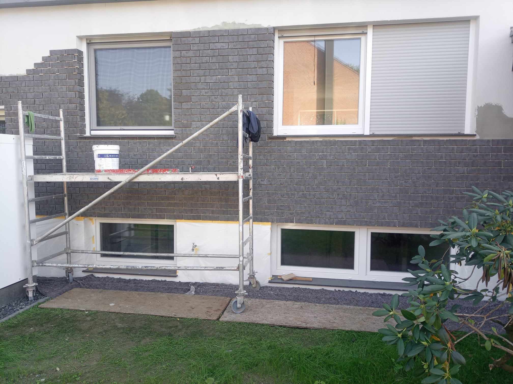 Montaż elewacji z szarej cegły klinkierowej na ścianie domu jednorodzinnego, widoczne rusztowanie, okna z roletami zewnętrznymi i fragment ogrodu z trawnikiem.