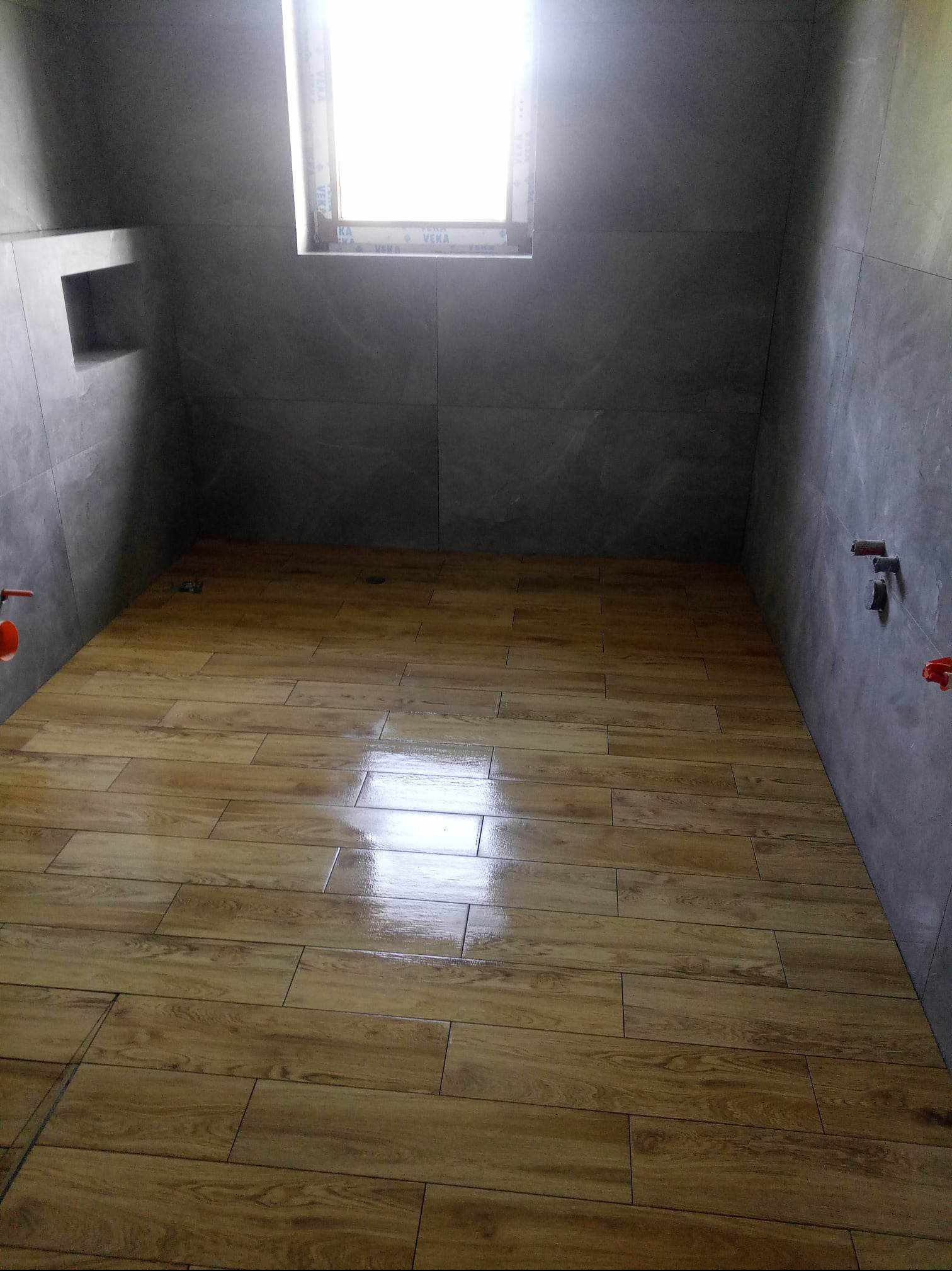 Wykończona łazienka z szarymi płytkami ściennymi i drewnopodobną podłogą z płytek, małe okno w górnej części ściany, widoczne elementy instalacji wodnej.