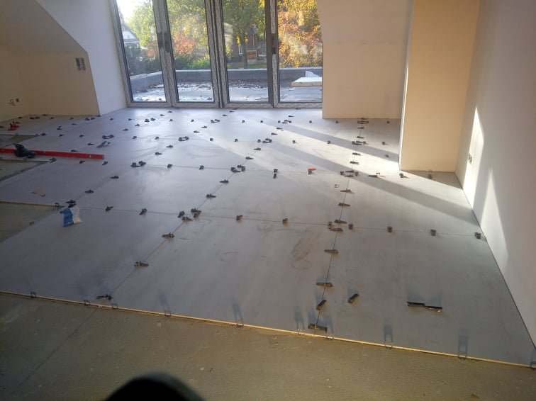 Szare płytki podłogowe ułożone z użyciem klipsów poziomujących w jasnym pomieszczeniu z dużymi oknami balkonowymi, widoczne narzędzia glazurnicze.