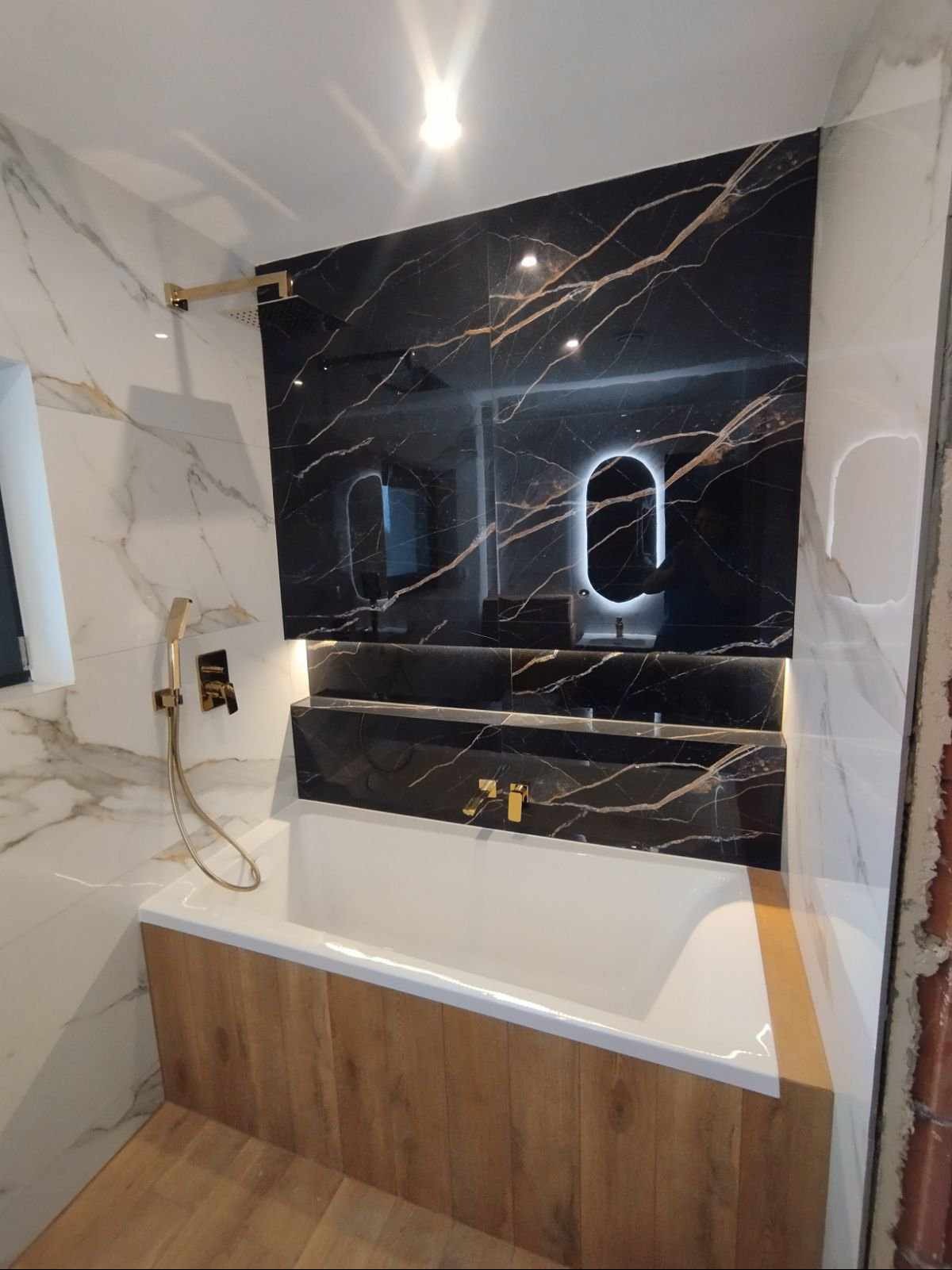 Nowoczesna łazienka z wanną obudowaną drewnopodobnymi panelami, czarne płytki z marmurowym wzorem na ścianie z podświetlanym lustrem i złotą armaturą.