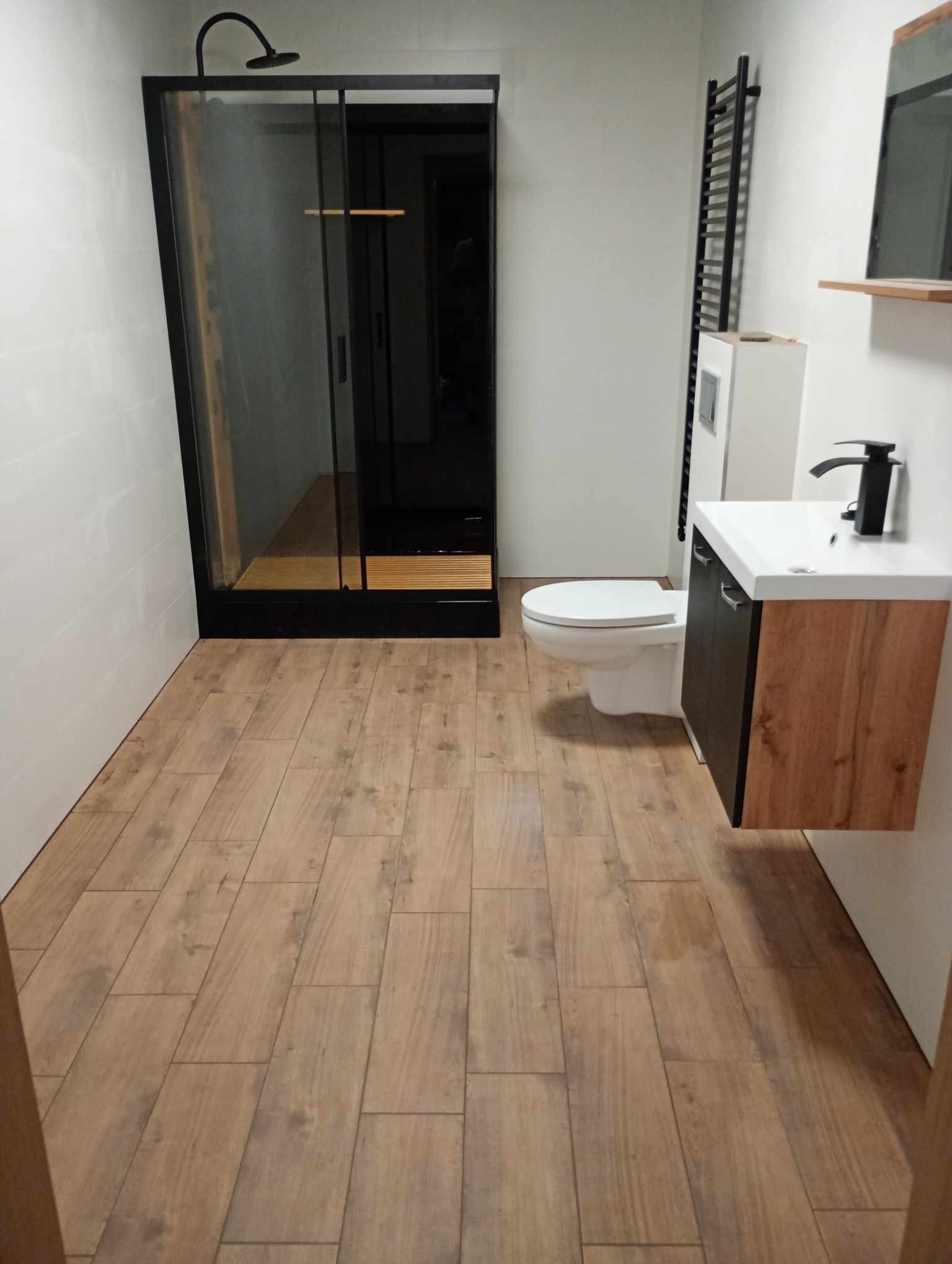 Nowoczesna łazienka z kabiną prysznicową typu walk-in z czarnymi ramami, umywalką z szafką w kolorze drewna i brązowymi płytkami podłogowymi imitującymi drewno.