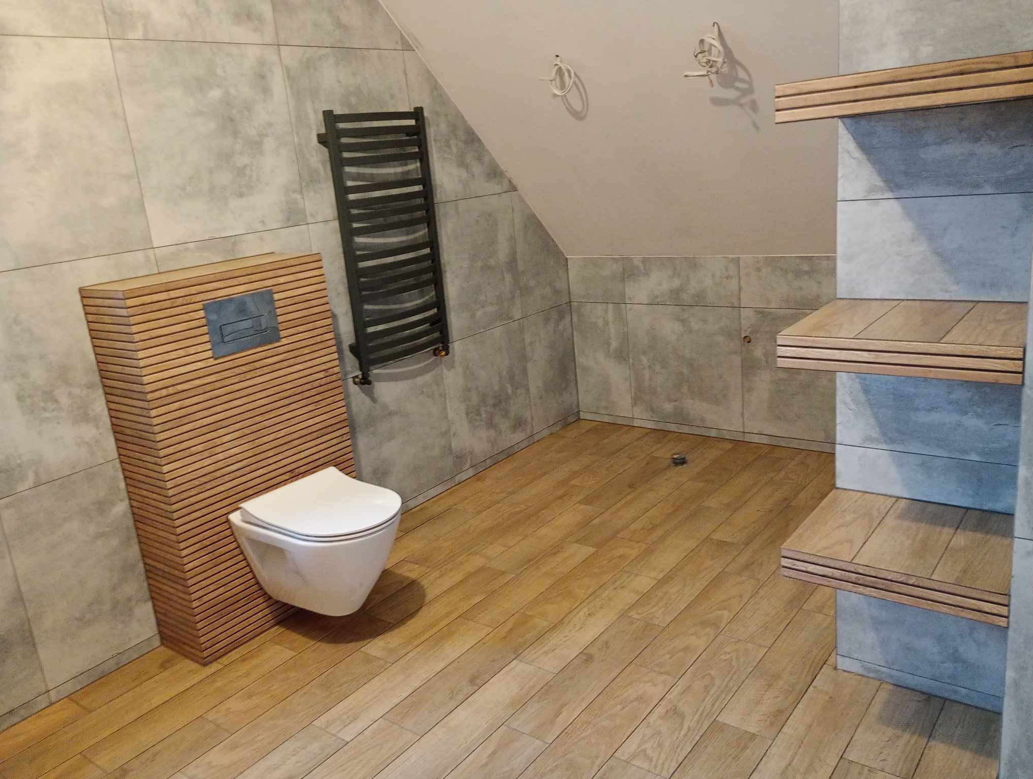 Nowoczesna łazienka na poddaszu z drewnopodobną podłogą, szarą płytką imitującą beton, podwieszaną toaletą z obudową z drewnianych listewek, grzejnikiem drabinkowym i drewnianymi półkami. Widoczne...