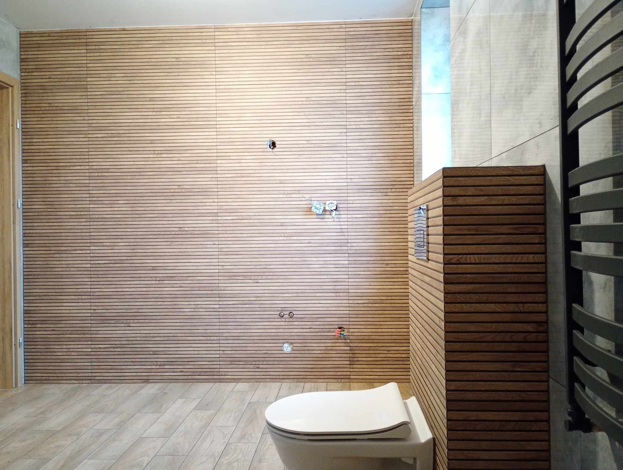 Łazienka w trakcie prac wykończeniowych: ściana wyłożona panelami imitującymi drewno z widocznymi puszkami elektrycznymi i rurami, podłoga z płytek w odcieniach beżu, częściowo zamontowana toaleta,...