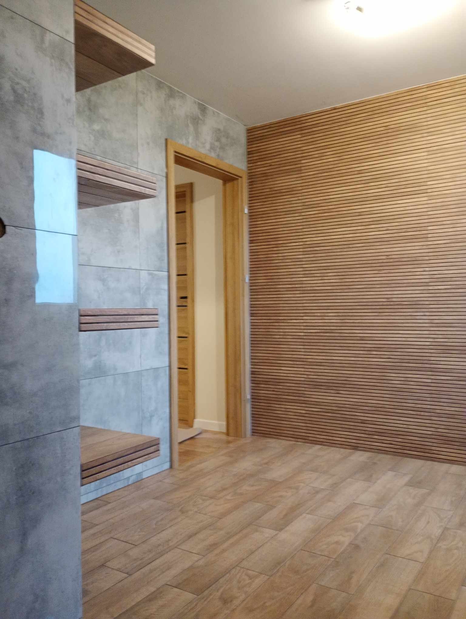 Wykończone wnętrze: ściana z drewnianych paneli, szara ściana z półkami z drewnianych listew i drewniana podłoga imitująca deski.