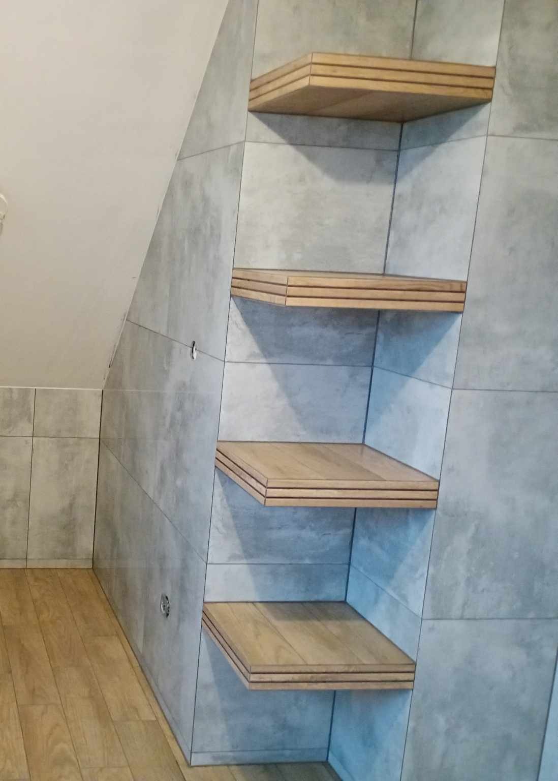 Wykończona łazienka na poddaszu z szarymi płytkami imitującymi beton i drewnianymi półkami wbudowanymi we wnękę