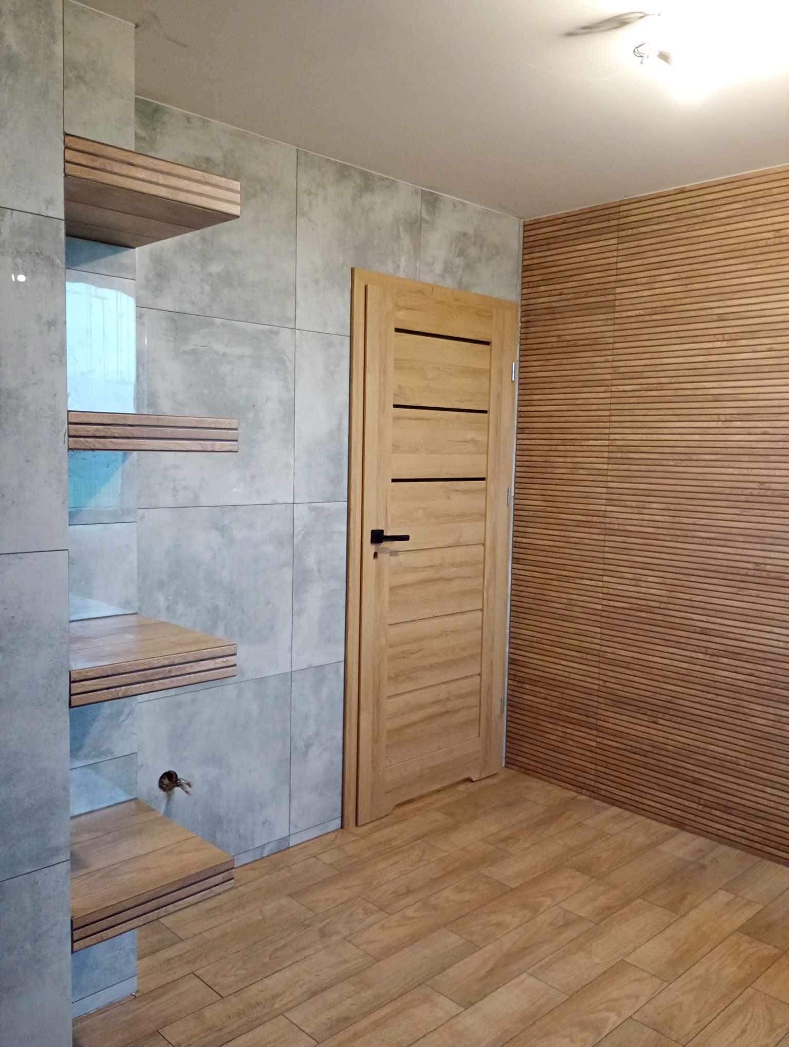 Wykończone wnętrze: szare płytki imitujące beton na ścianie z wbudowanymi drewnianymi półkami, drewniane panele ścienne w odcieniach brązu oraz jasne drewniane drzwi z poziomymi frezowaniami.