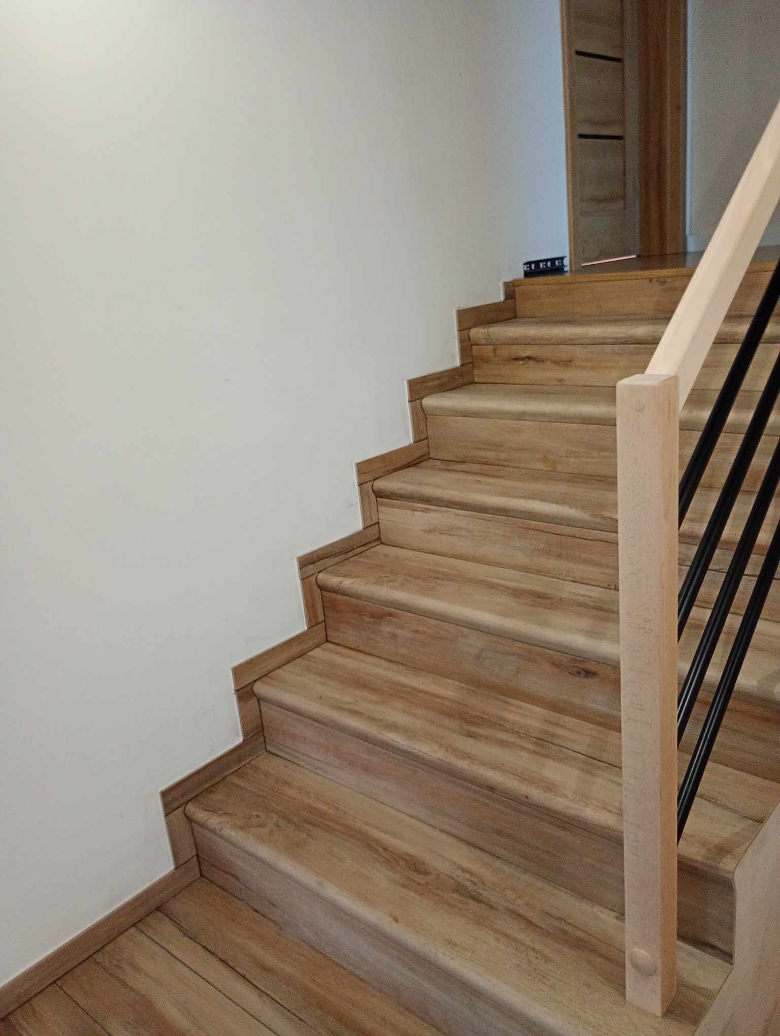 Schody drewniane z jasną balustradą i czarnymi prętami, wykończone listwami przypodłogowymi w kolorze drewna, widok z dołu.