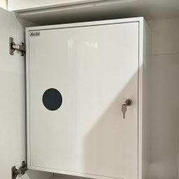 Tomodom Tomasz Turski - Biała skrzynka elektryczna z okrągłym otworem wentylacyjnym i kluczem w zamku, umieszczona w szafce, obok widoczne bezpieczniki topikowe.