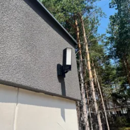 Czarna, nowoczesna lampa elewacyjna zamontowana na szarej, teksturowanej ścianie domu, w tle wysokie sosny i błękitne niebo.