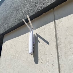 Tomodom Tomasz Turski - Biały, dwuantenowy punkt dostępowy WiFi zamontowany na teksturowanej, jasnej ścianie budynku w słoneczny dzień, rzucający wyraźny cień.