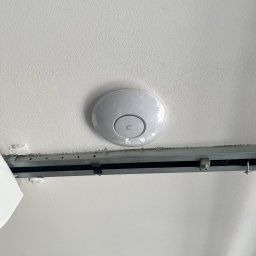 Tomodom Tomasz Turski - Biały, okrągły punkt dostępowy Wi-Fi zamontowany na suficie podwieszanym z metalową szyną montażową.