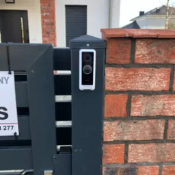 Nowoczesny wideofon w białej obudowie zamontowany na grafitowym słupku ogrodzenia z ceglanym fragmentem muru w tle.