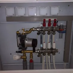 Rozdzielacz ogrzewania podłogowego w szafce z widocznymi etykietami pomieszczeń: łazienka, korytarz, sypialnia, biuro. Widoczny manometr i pompa obiegowa Upornor.