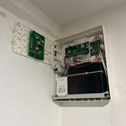 Tomodom Tomasz Turski - Otwarta, biała skrzynka alarmowa z elektroniką, akumulatorem i przewodami zamontowana na białej ścianie w rogu pomieszczenia. Widoczne płytki drukowane i bateria.
