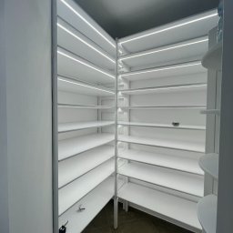 Tomodom Tomasz Turski - Biała, pusta garderoba z półkami oświetlonymi listwami LED, widok z dołu, z elementami instalacji elektrycznej na półkach. Minimalistyczny design.