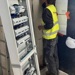Tomodom Tomasz Turski - Otwarta, biała skrzynka rozdzielcza z licznymi bezpiecznikami i niebieskimi przewodami. Elektryk w żółtej kamizelce montuje instalację w surowym wnętrzu.