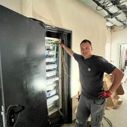 Tomodom Tomasz Turski - Elektryk w czerwonych rękawicach prezentuje rozdzielnicę elektryczną w trakcie montażu w nowym budynku. Widoczne kable i elementy instalacji.