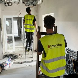 Tomodom Tomasz Turski - Prace instalacyjne w Zubrzycy Górnej: Dwóch monterów w kamizelkach 'tomodom' na drabinach, instalacje pod sufitem, przewody na podłodze, remont w toku.