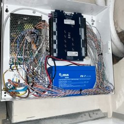 Tomodom Tomasz Turski - Rozbudowana instalacja alarmowa w białej obudowie, z akumulatorem Zeus FX-7 i licznymi przewodami. Widoczny kontroler dostępu KaDe.