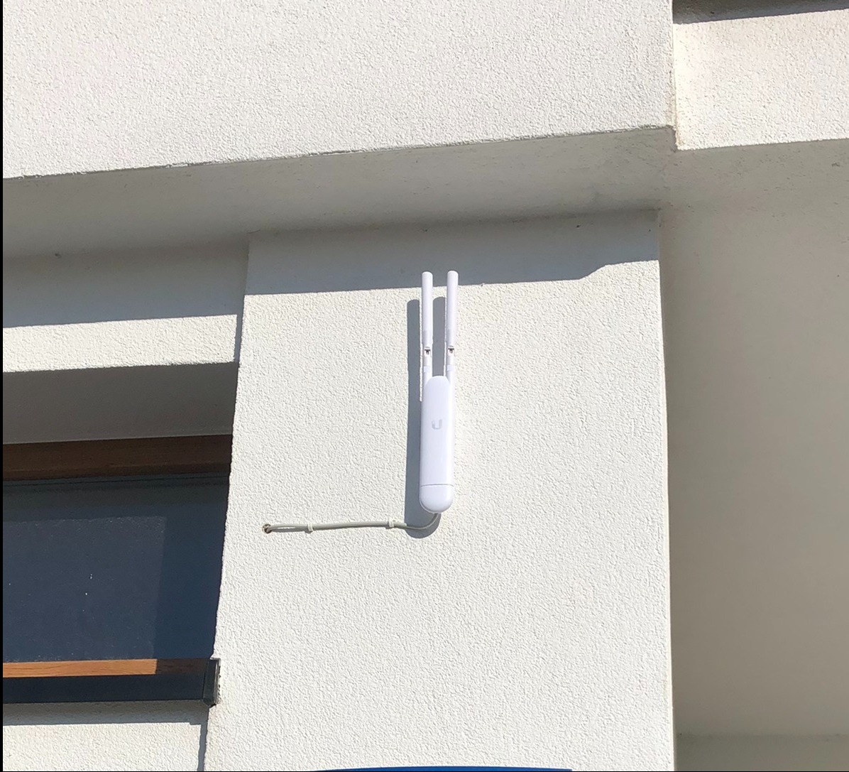 Biały punkt dostępowy WiFi, model Ubiquiti, zamontowany na elewacji budynku obok okna, z widocznym kablem zasilającym.
