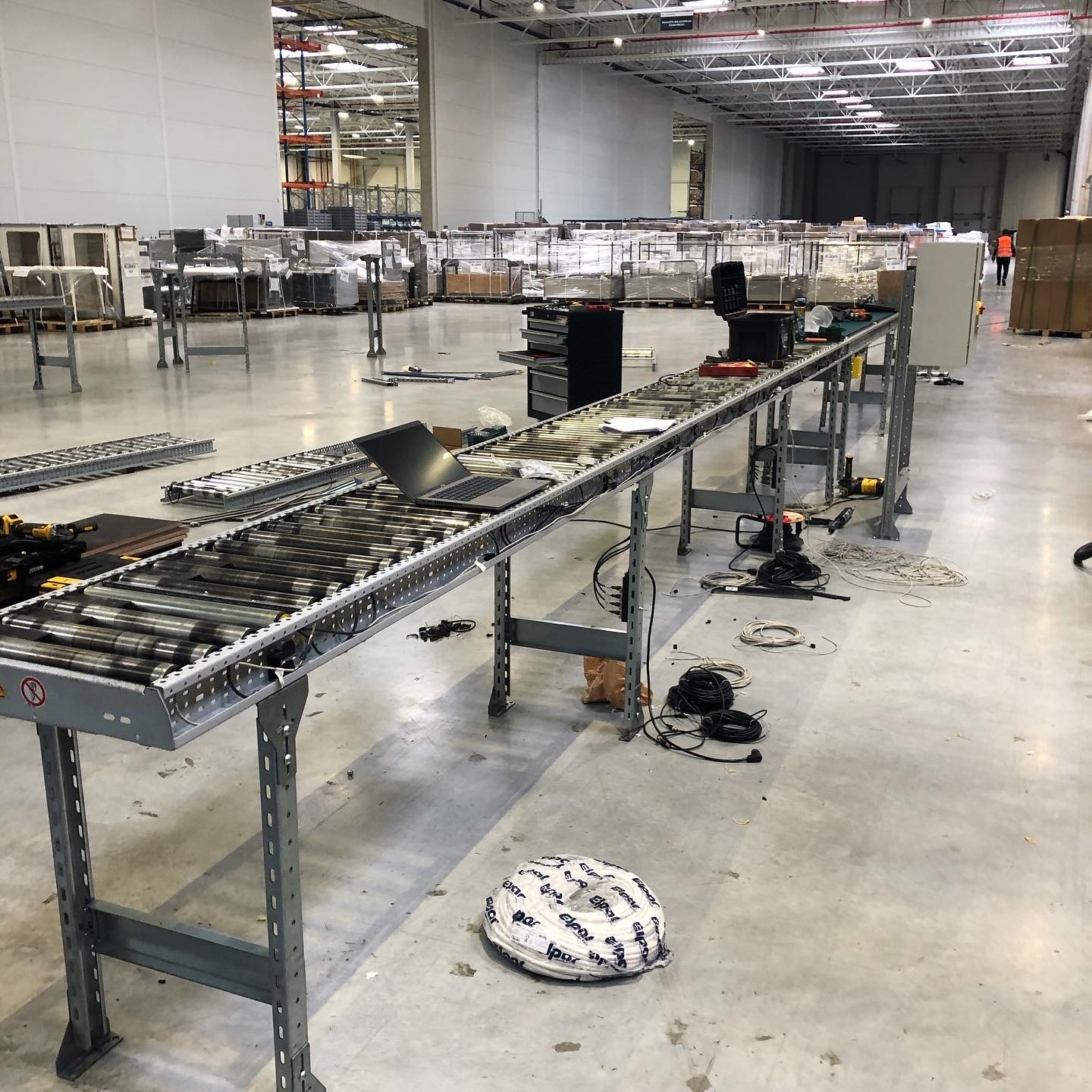 Rozległa hala magazynowa z montowanym systemem transportu rolkowego; widoczny laptop na fragmencie taśmy, narzędzia, przewody elektryczne oraz zwój kabla na podłodze. W tle regały magazynowe...