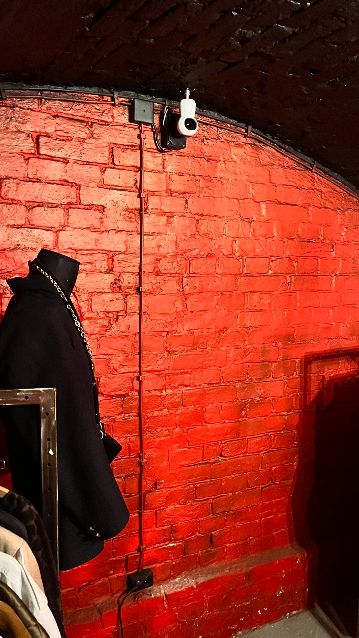 Biała kamera monitoringu zamontowana na czerwonej, ceglanej ścianie w pomieszczeniu z czarnym sufitem, widoczne przewody i fragment manekina z ubraniem.