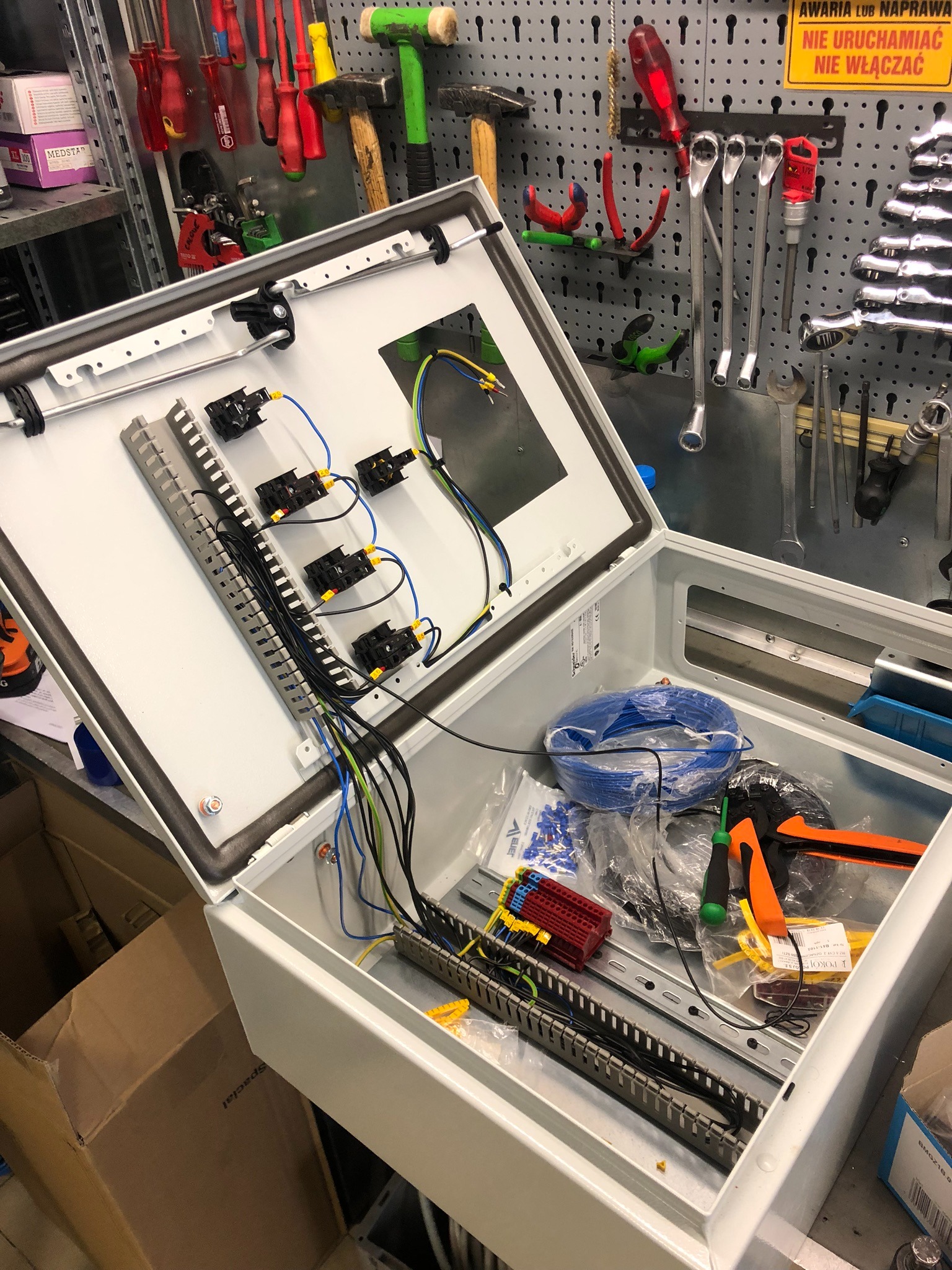 Otwarta, biała skrzynka elektryczna z zamontowanymi elementami i podłączonymi przewodami; widoczne listwy zaciskowe, przewody w kolorach niebieskim, żółtym i czarnym, oraz narzędzia w tle...