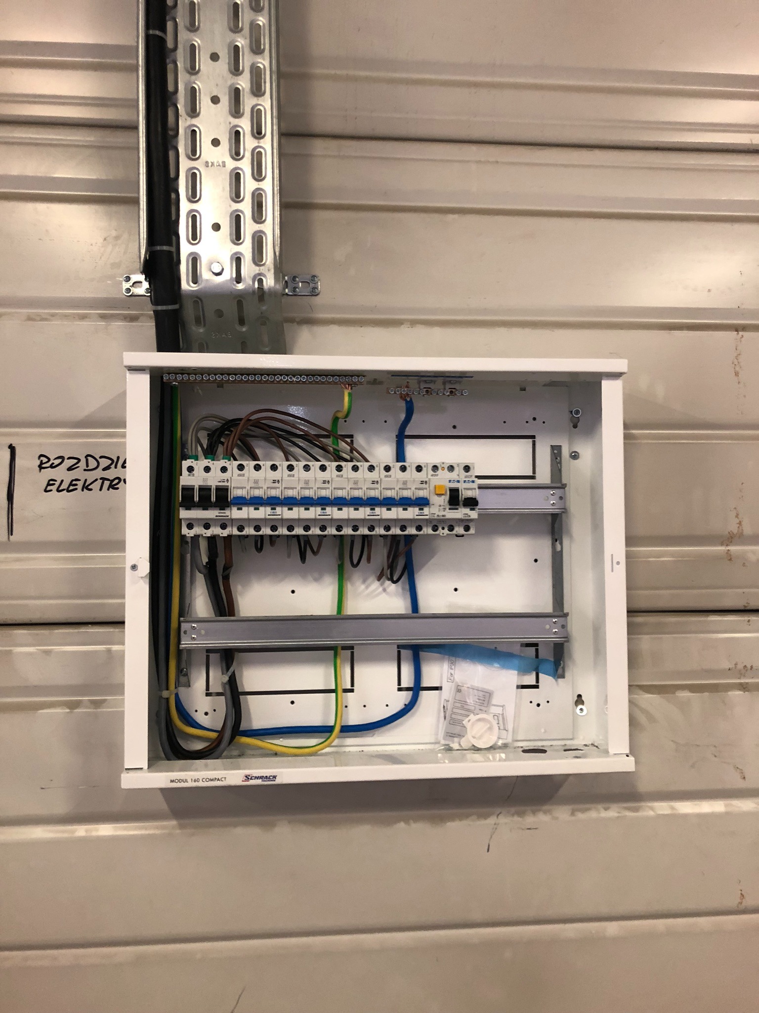 Otwarta, biała skrzynka rozdzielcza z zainstalowanymi bezpiecznikami i podłączonymi kablami, zamontowana na metalowej ścianie z widocznym napisem 'ROZDZIELNIA ELEKTRYCZNA' obok.