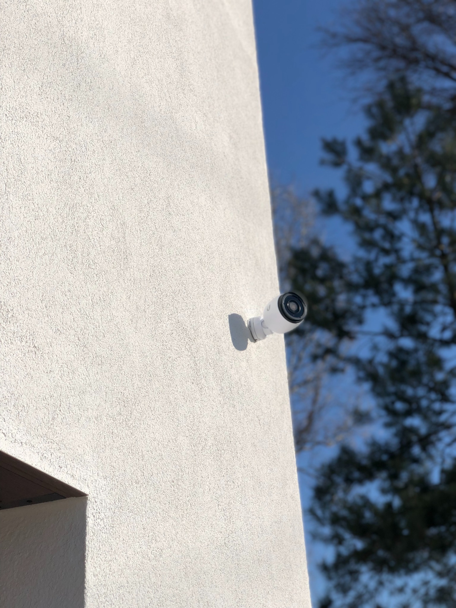 Biała kamera monitoringu przymocowana do chropowatej ściany budynku, na tle błękitnego nieba i gałęzi drzew.