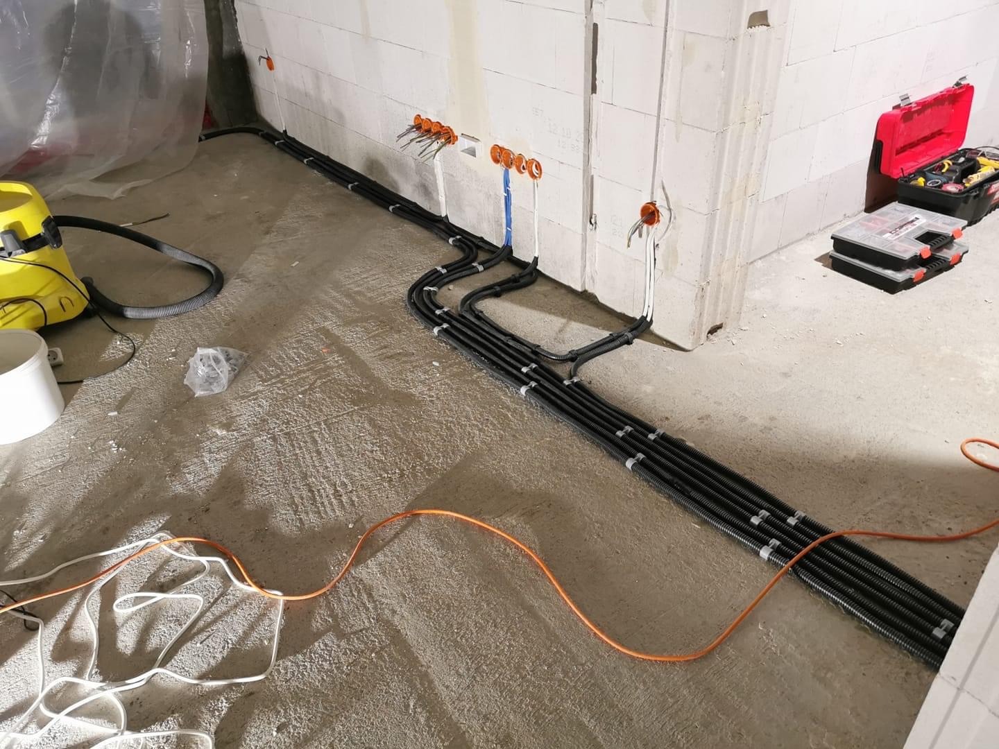 Układanie peszli z kablami elektrycznymi w surowym wnętrzu budynku z bloczków, widoczna skrzynka narzędziowa i odkurzacz przemysłowy.