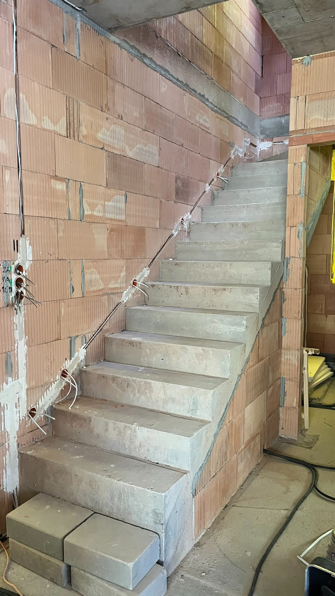 Surowe betonowe schody w trakcie budowy w Zubrzycy Górnej, z widocznymi przewodami elektrycznymi prowadzonymi wzdłuż ściany z cegły ceramicznej, puszki elektryczne z wystającymi przewodami.