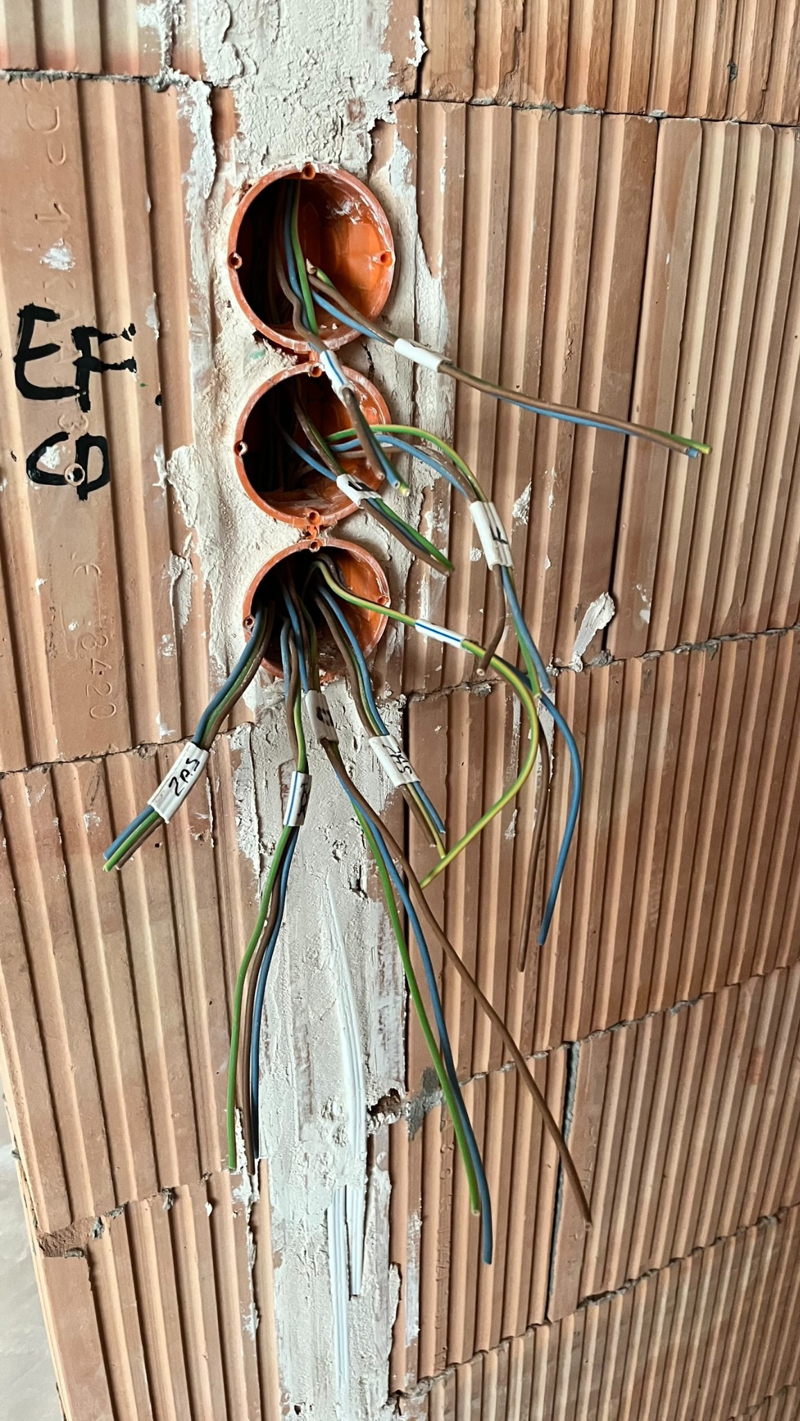 Trzy pomarańczowe puszki elektryczne osadzone w murze z cegły ceramicznej, wystające przewody elektryczne oznaczone etykietami, świeże ślady zaprawy murarskiej.