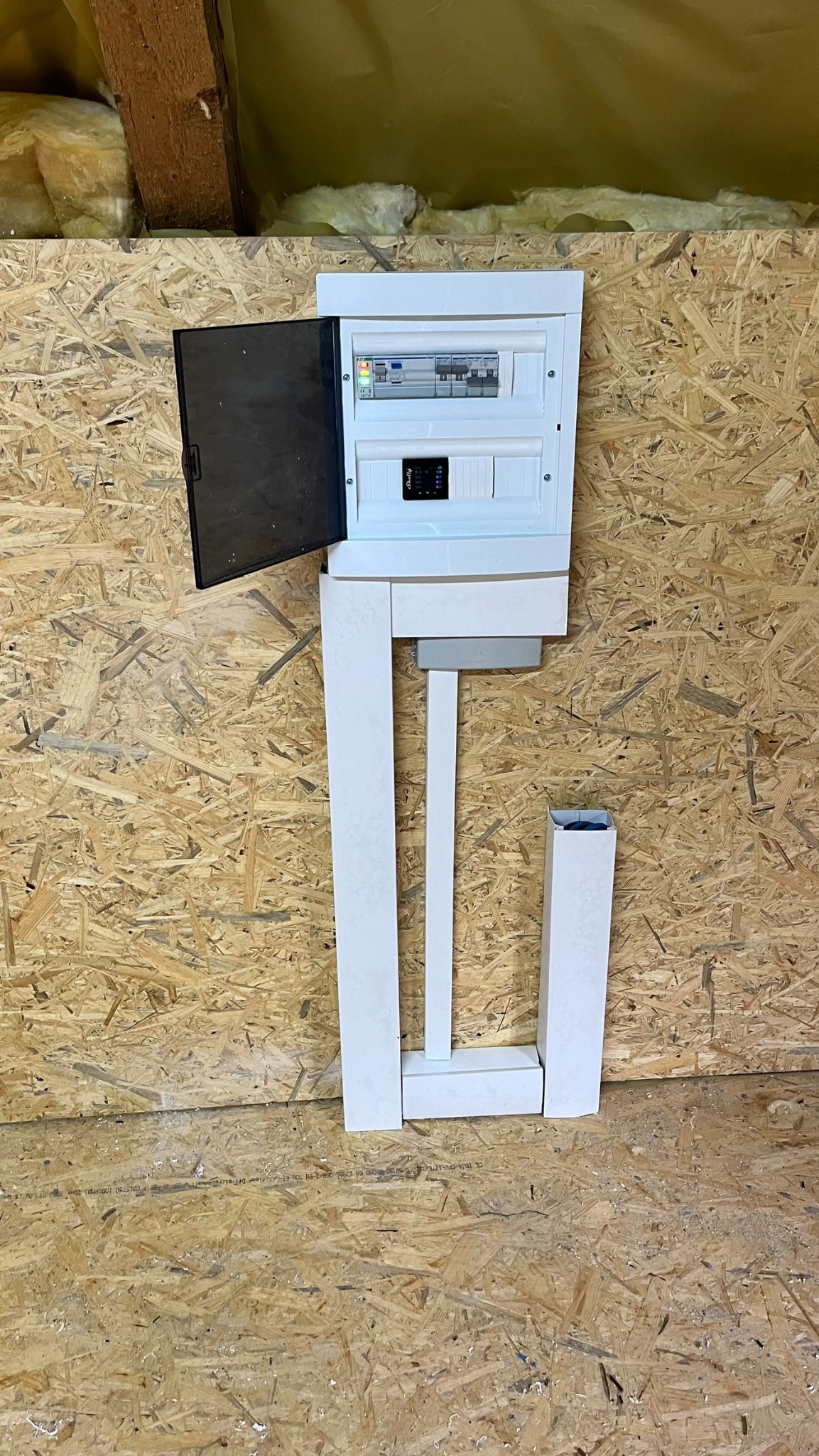 Rozdzielnica elektryczna z otwartymi drzwiczkami, zamontowana na ścianie z płyty OSB, widoczne przewody w białych maskownicach.