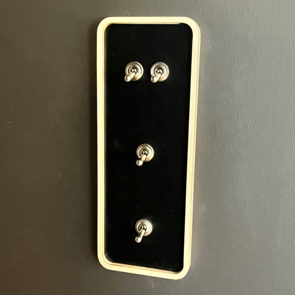 Czarny panel z trzema chromowanymi, przełącznikami dźwigniowymi, otoczony jasną, drewnianą ramką, zamontowany na szarej ścianie.