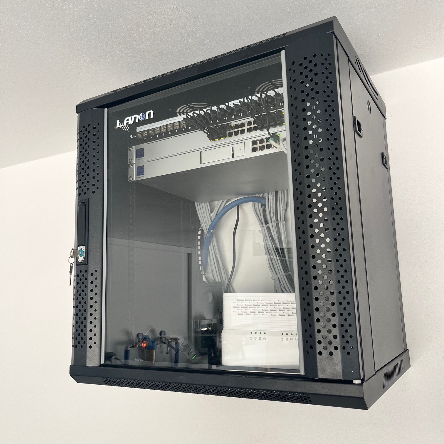 Czarna, wisząca szafka rack Lanon z przeszklonymi drzwiami, zawierająca router, switche i okablowanie, zamontowana na białej ścianie.