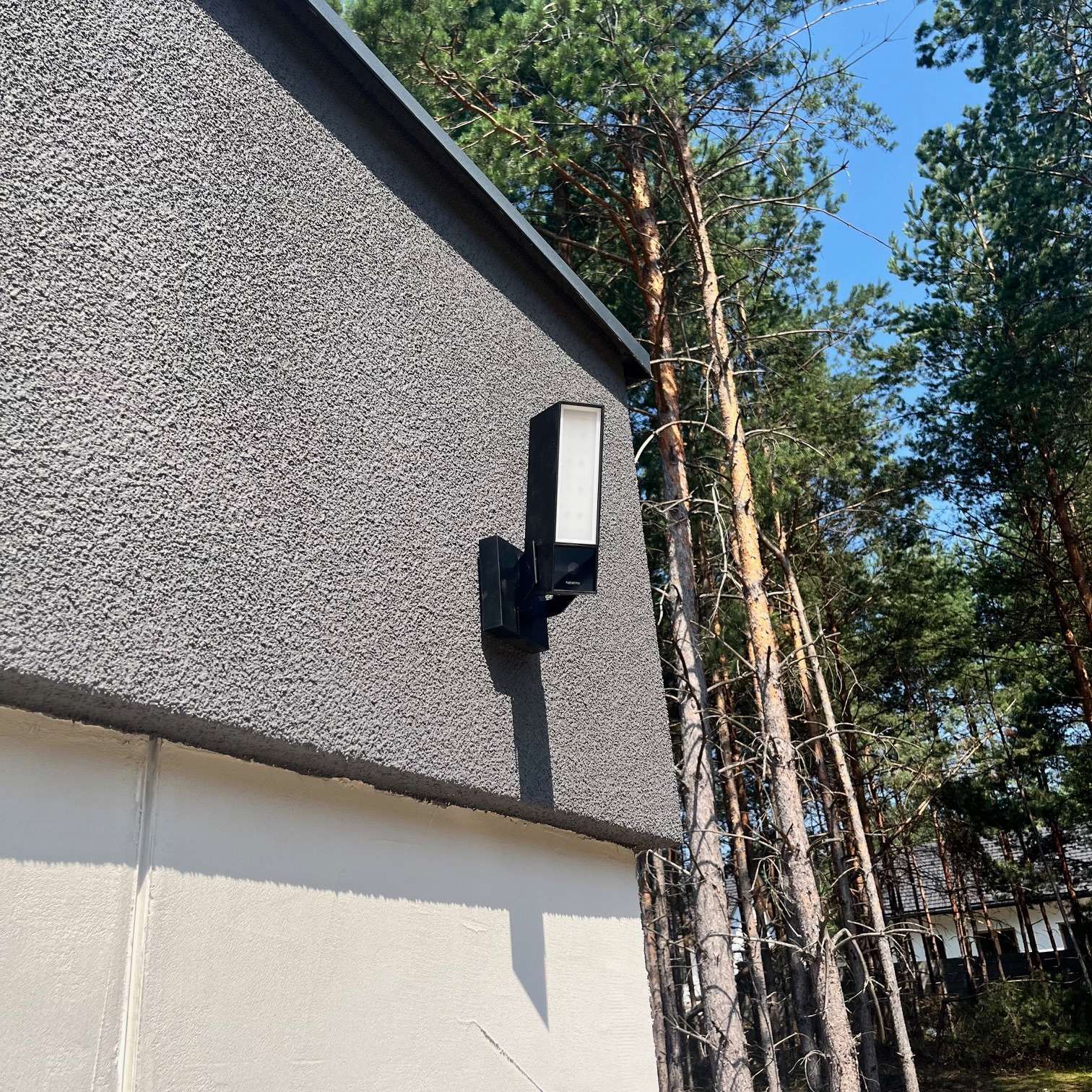 Czarna, nowoczesna lampa elewacyjna zamontowana na szarej, teksturowanej ścianie domu, w tle wysokie sosny i błękitne niebo.