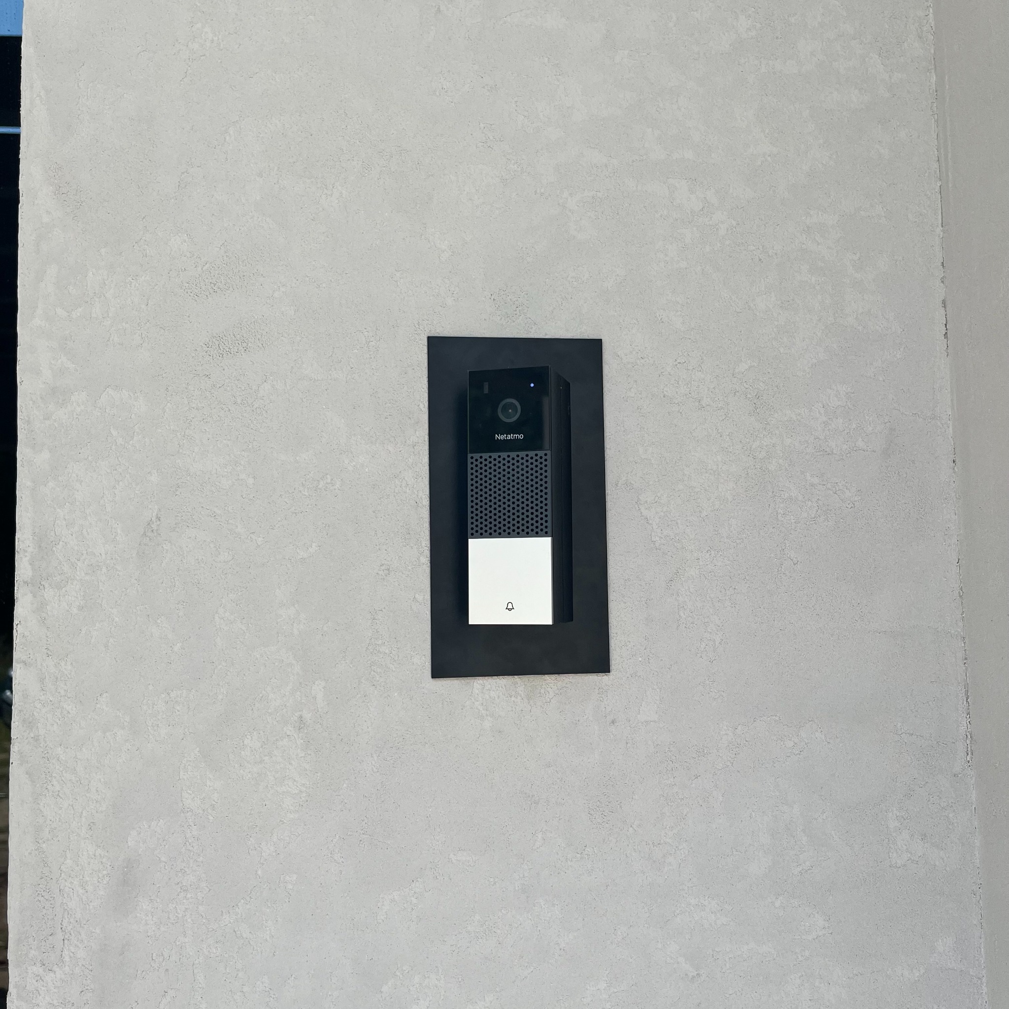 Zewnętrzny wideodomofon Netatmo zamontowany na szarej, teksturowanej ścianie, otoczony czarną ramką.
