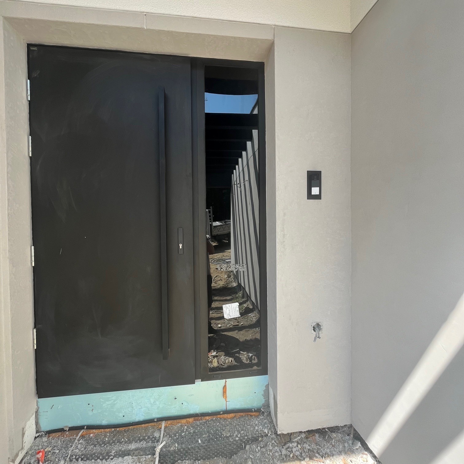 Nowoczesne, matowe, grafitowe drzwi wejściowe z wąskim oknem bocznym i minimalistycznym panelem domofonowym na jasnej, teksturowanej ścianie. W tle widoczne elementy konstrukcji budynku w trakcie...