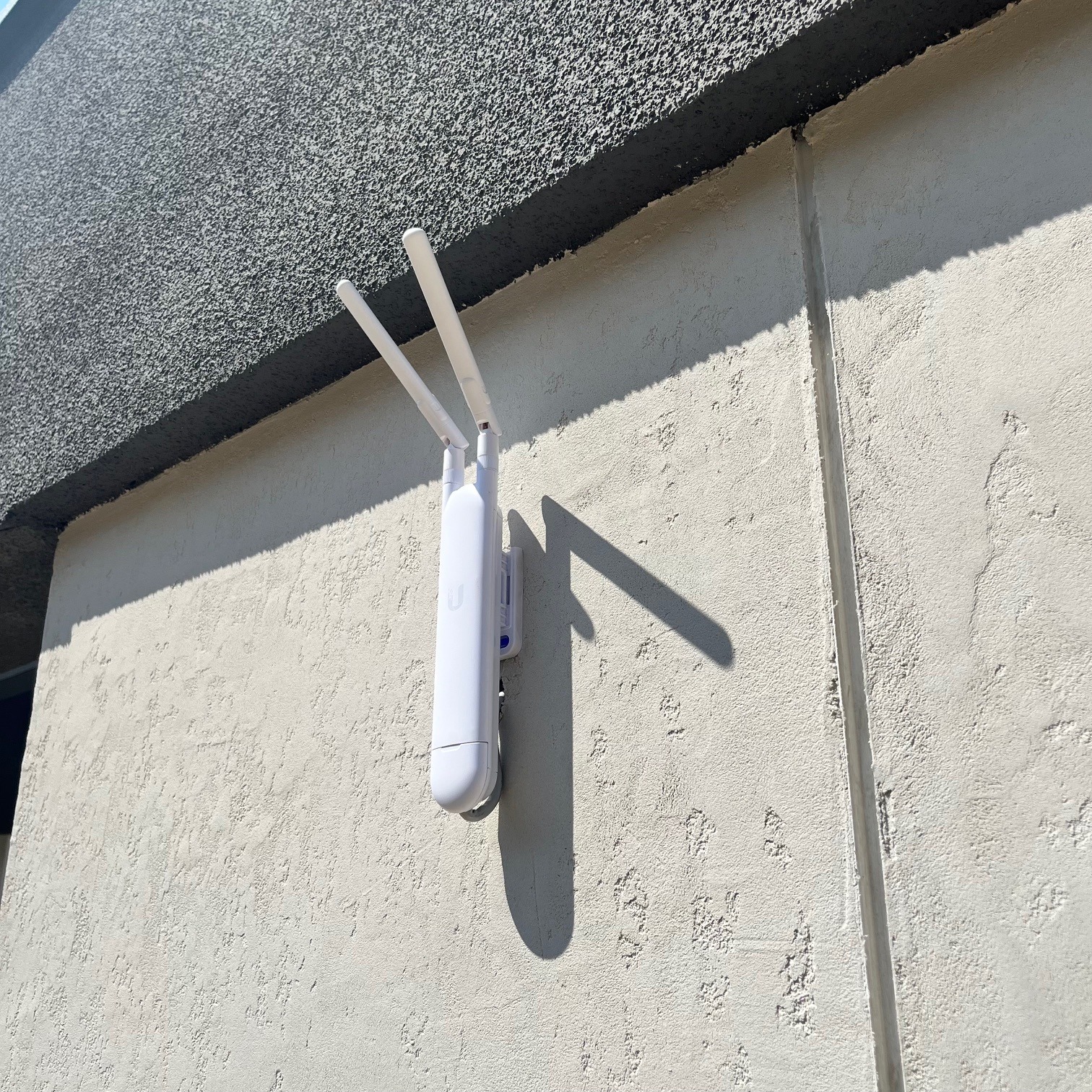 Biały, dwuantenowy punkt dostępowy WiFi zamontowany na teksturowanej, jasnej ścianie budynku w słoneczny dzień, rzucający wyraźny cień.