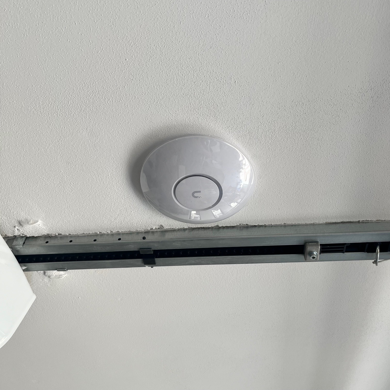 Biały, okrągły punkt dostępowy Wi-Fi zamontowany na suficie podwieszanym z metalową szyną montażową.