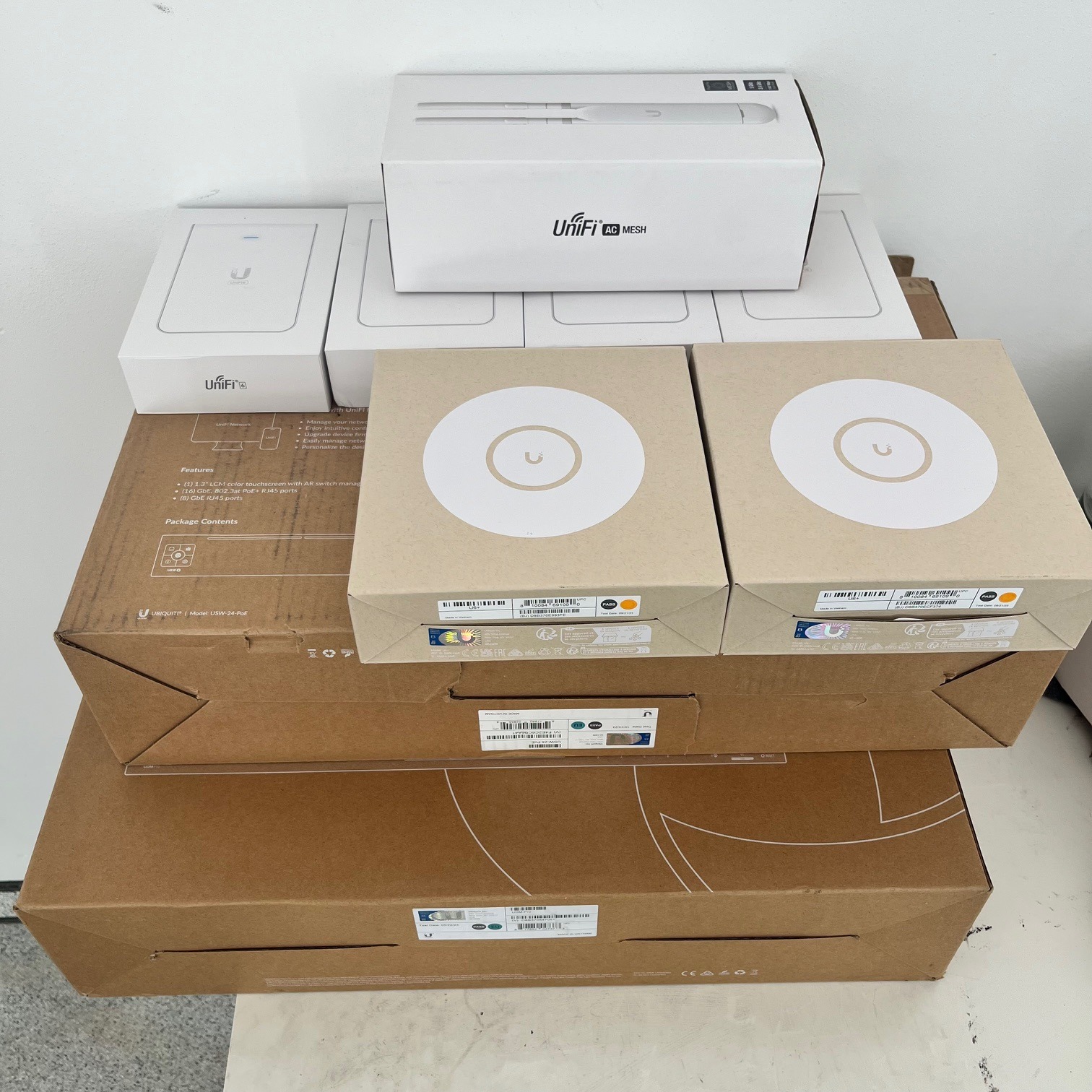Stos pudełek z urządzeniami sieciowymi Unifi, w tym routery i punkty dostępowe, w oryginalnych opakowaniach, ułożonych na sobie przed instalacją.