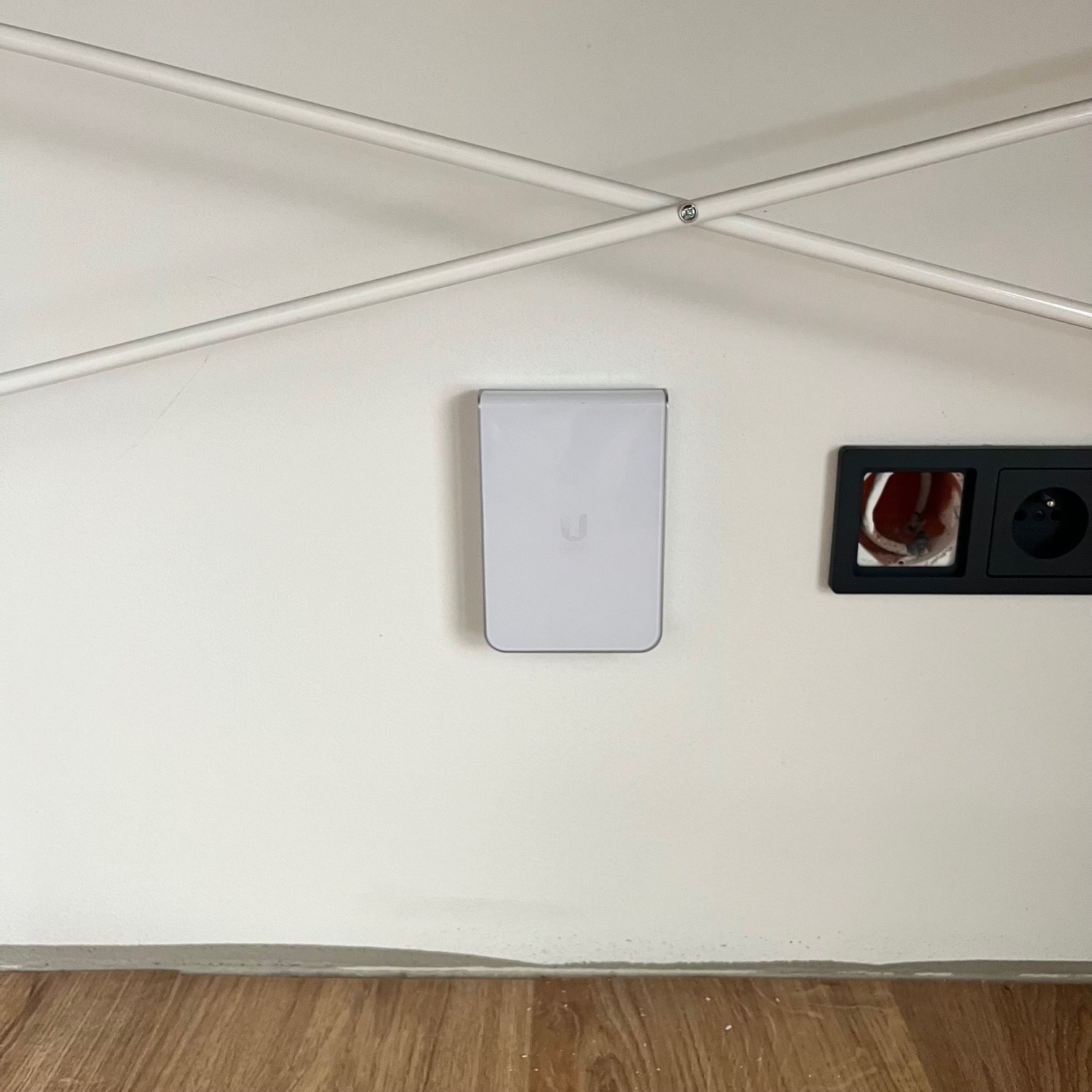 Biała ściana z zamontowanym punktem dostępowym WiFi (Ubiquiti) obok gniazdek elektrycznych w czarnej ramce, widoczna konstrukcja sufitowa z białych rurek.