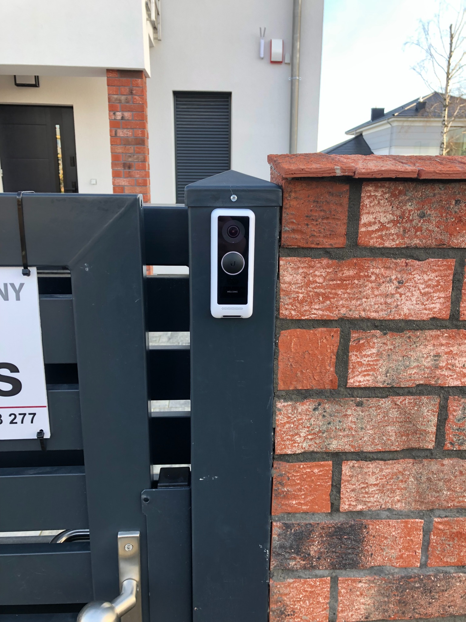 Nowoczesny wideofon w białej obudowie zamontowany na grafitowym słupku ogrodzenia z ceglanym fragmentem muru w tle.