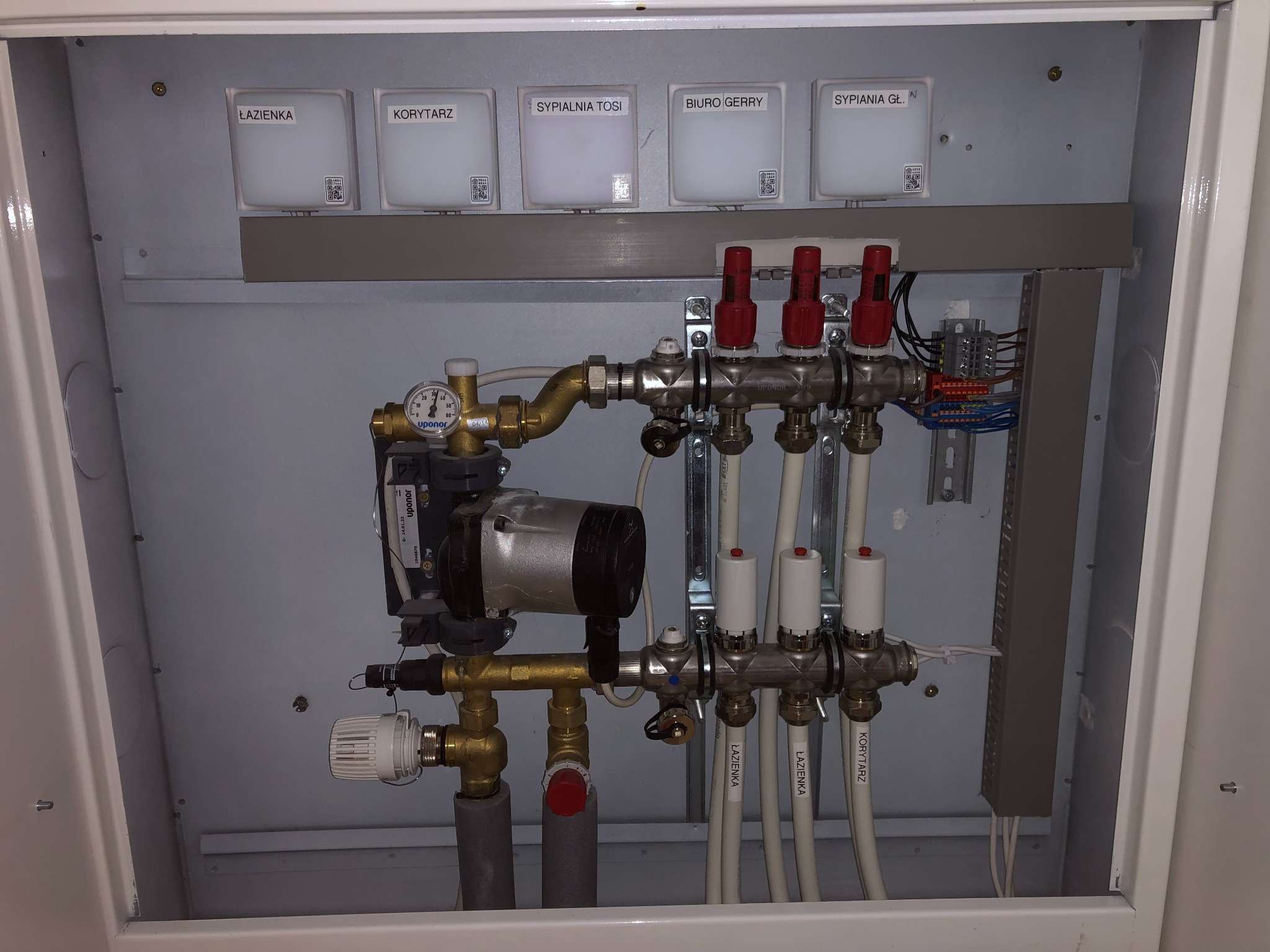 Rozdzielacz ogrzewania podłogowego w szafce z widocznymi etykietami pomieszczeń: łazienka, korytarz, sypialnia, biuro. Widoczny manometr i pompa obiegowa Upornor.