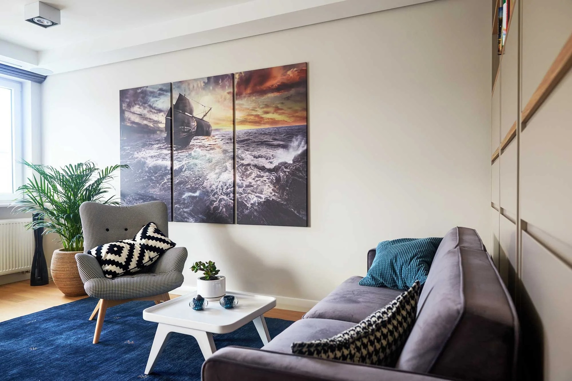 Salon z szarą sofą, fotel w pepitkę, dywan w odcieniach niebieskiego i obrazem przedstawiającym żaglowiec na wzburzonym morzu, podzielonym na trzy części.